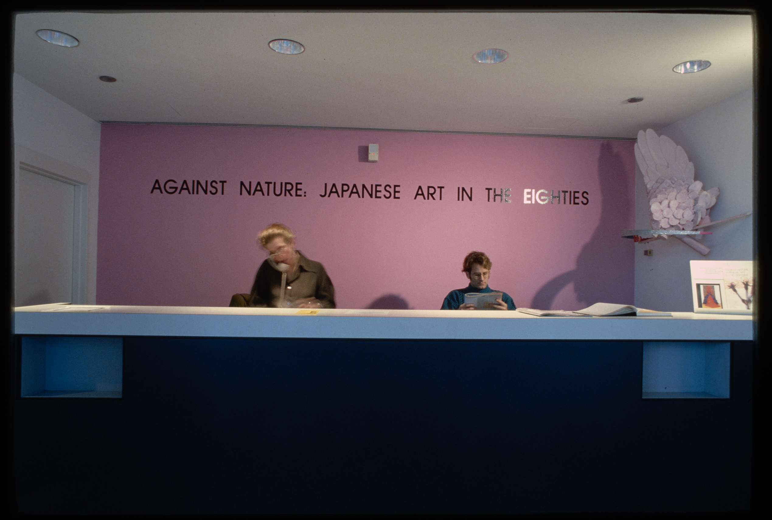 2 people sit at a desk in front of a bright pink wall with the exhibit name in black, a shelf on the right holds a sculpture.
