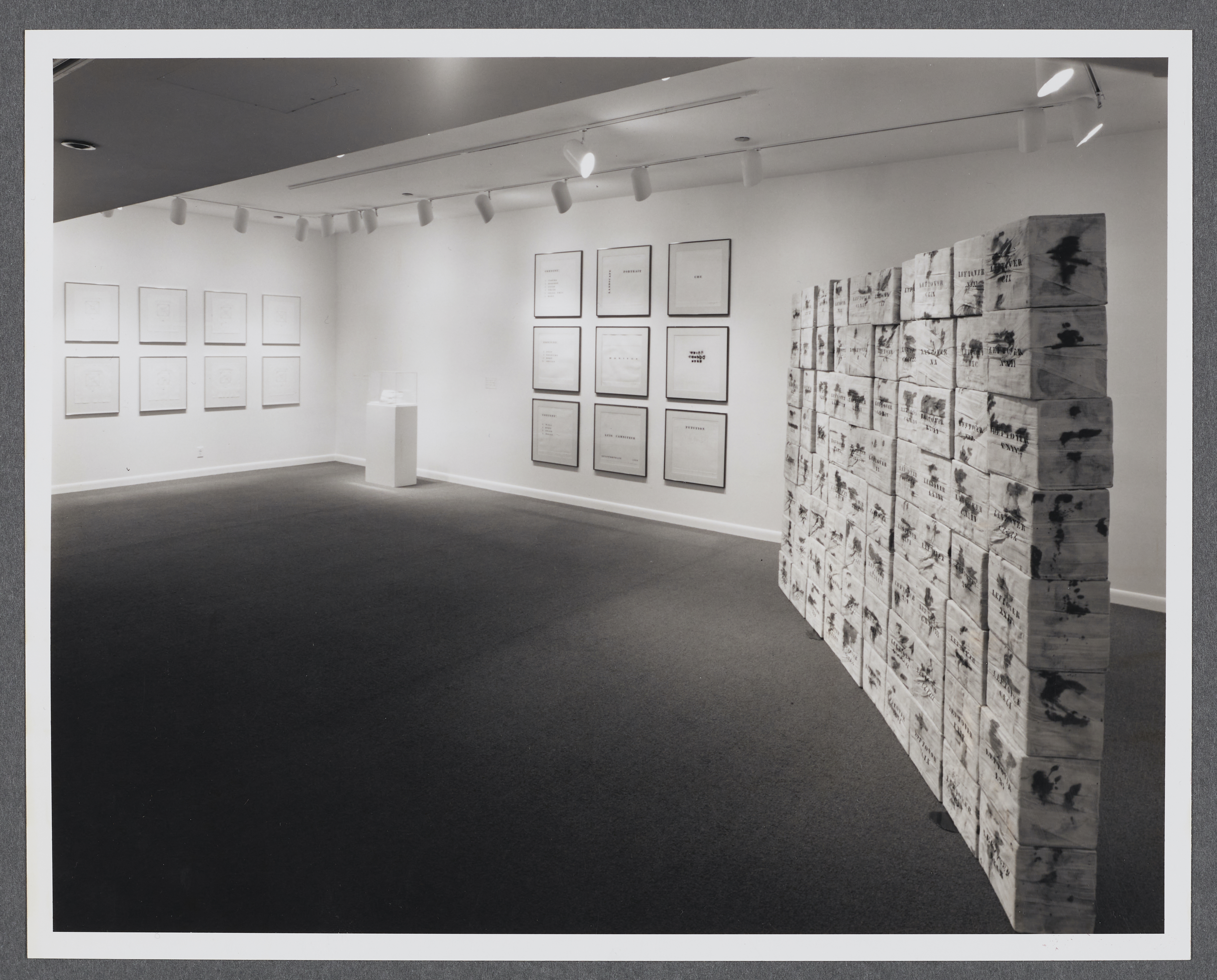 Framed works are hung in grids on the walls. A small pedestal sits in the corner while a wall sculpture sits on the opposite side.