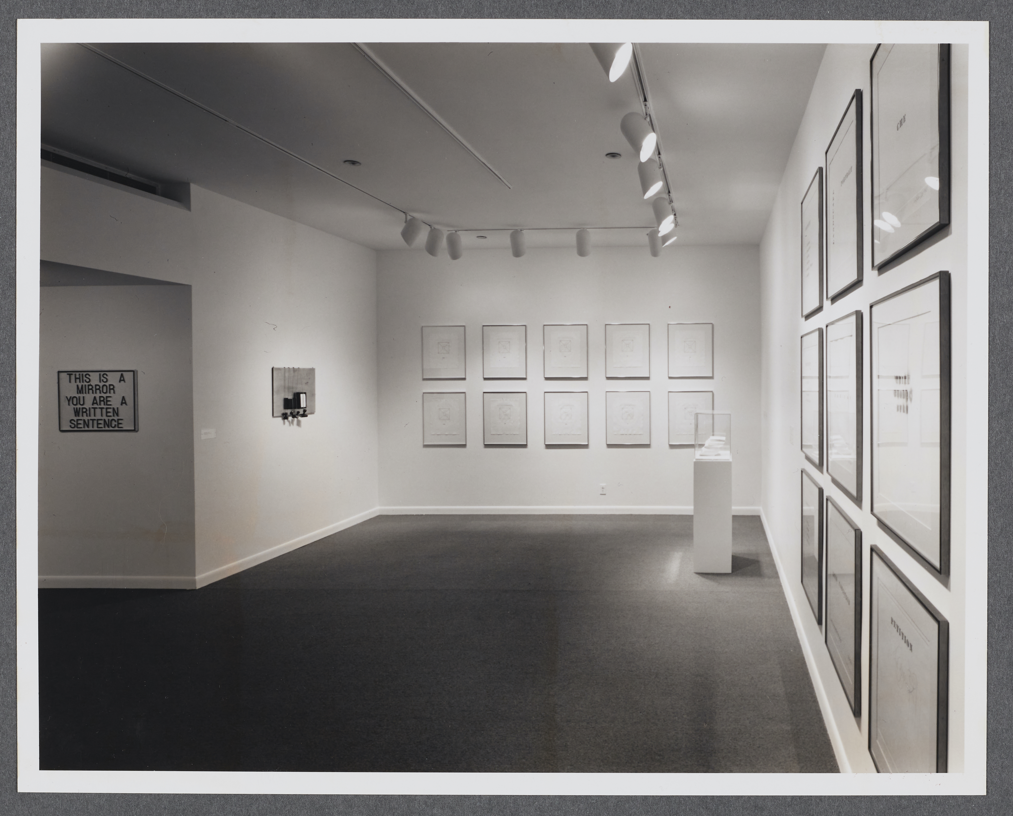 Framed works line the gallery wall in different configurations. A pedestal holding a small sculpture sits in the corner.