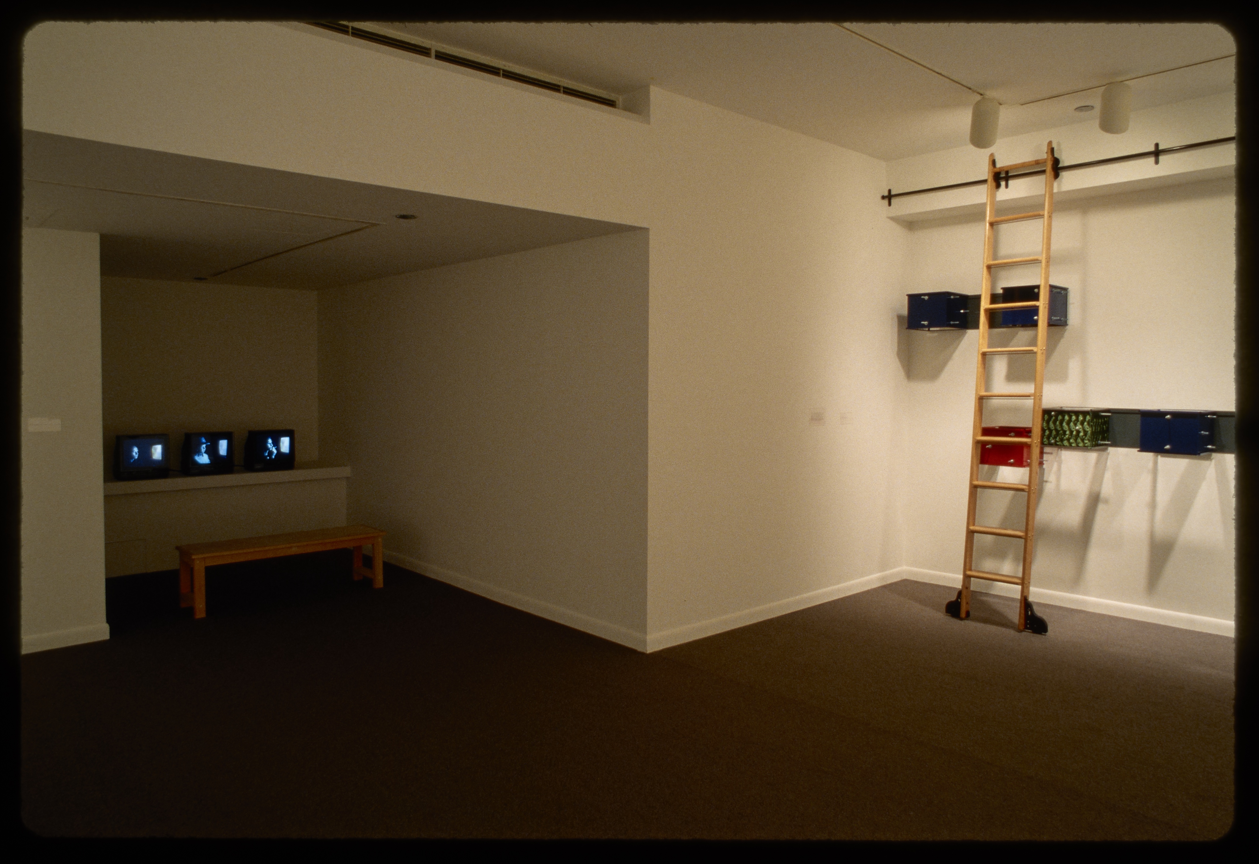 Bench sits in front of three videos playing on small tvs in alcove, on adjacent wall ladder stands in front of colorful boxes.