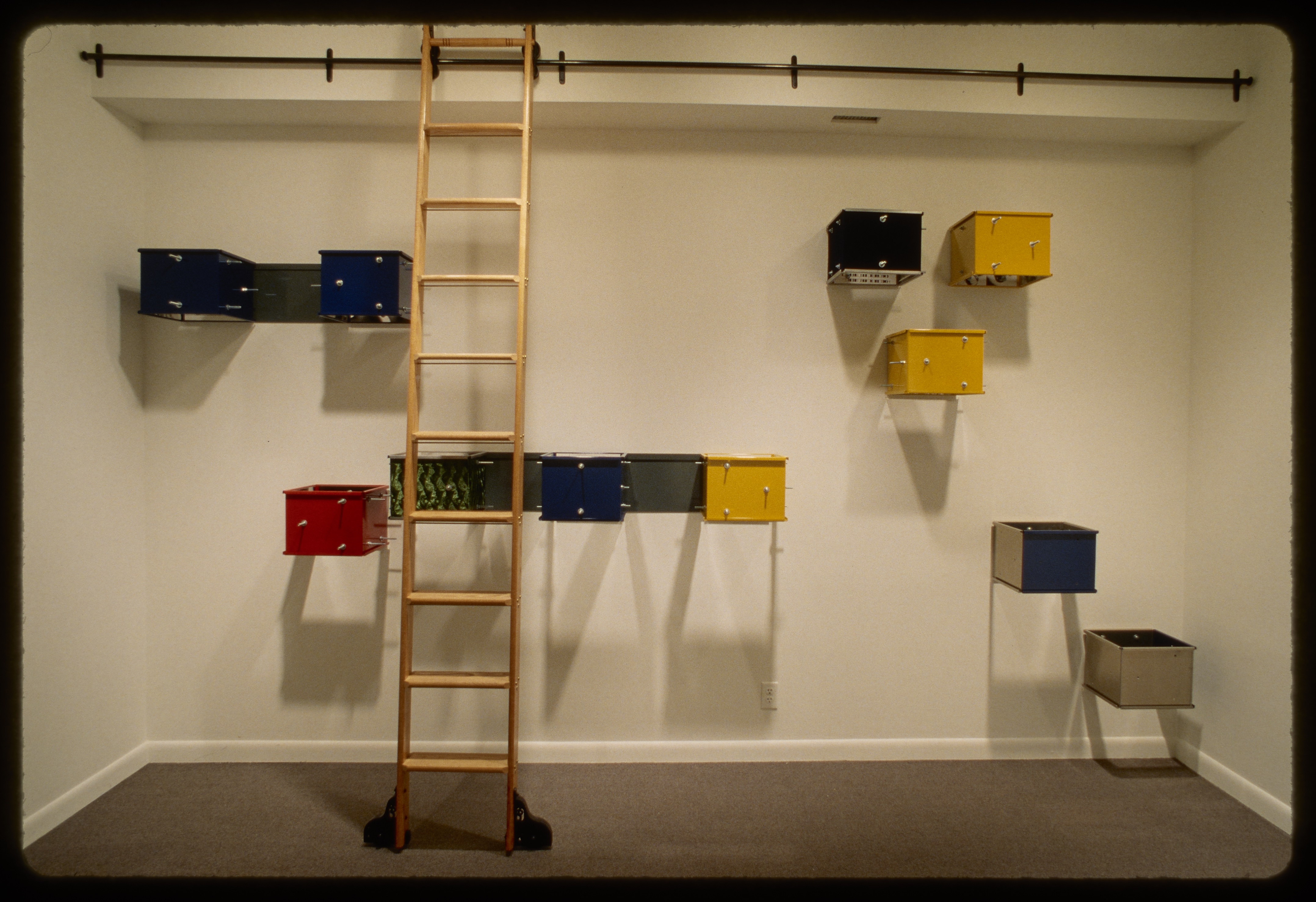 Wooden ladder stands in front of blue, yellow, red, and green metal boxes hung in asymmetric groups on the gallery wall.