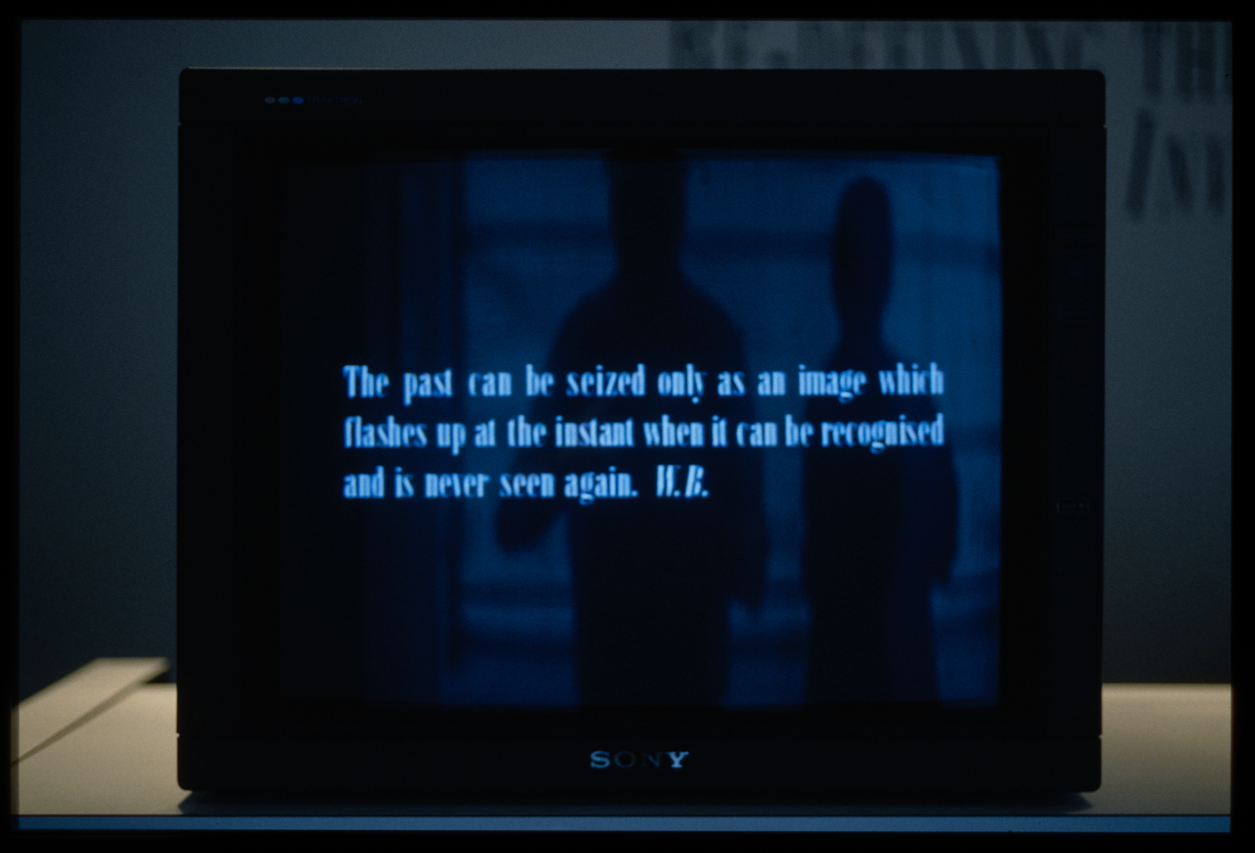 A monitor sits on a dimly lit surface, displaying light blue text on top of 2 shadow figures on a dark blue background.