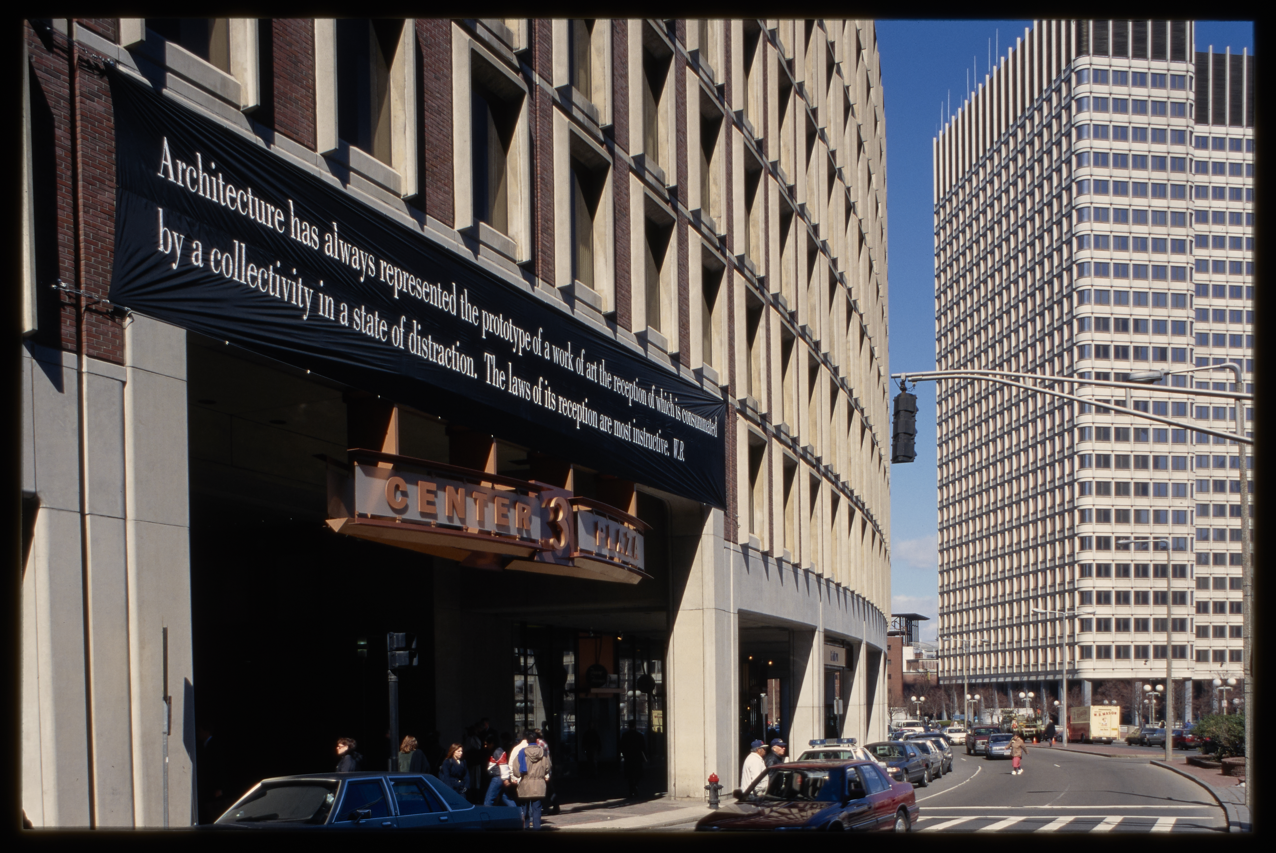 Seen at an angle, a black banner with white text is displayed above the entry of a large curved building on a city street.