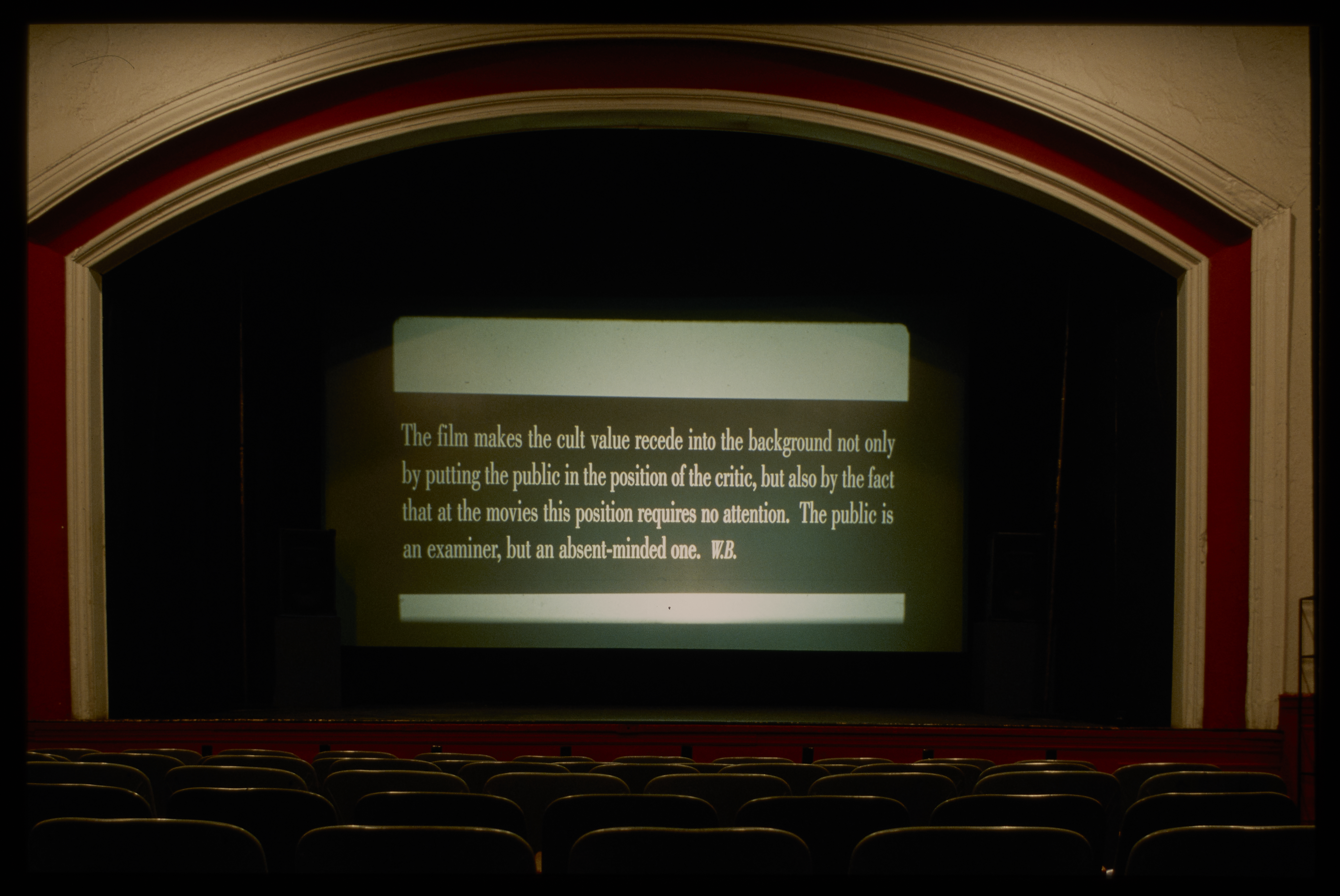 In an unoccupied theater, in the center of the stage, a screen displays a quote on a dark banner, over a blank background.