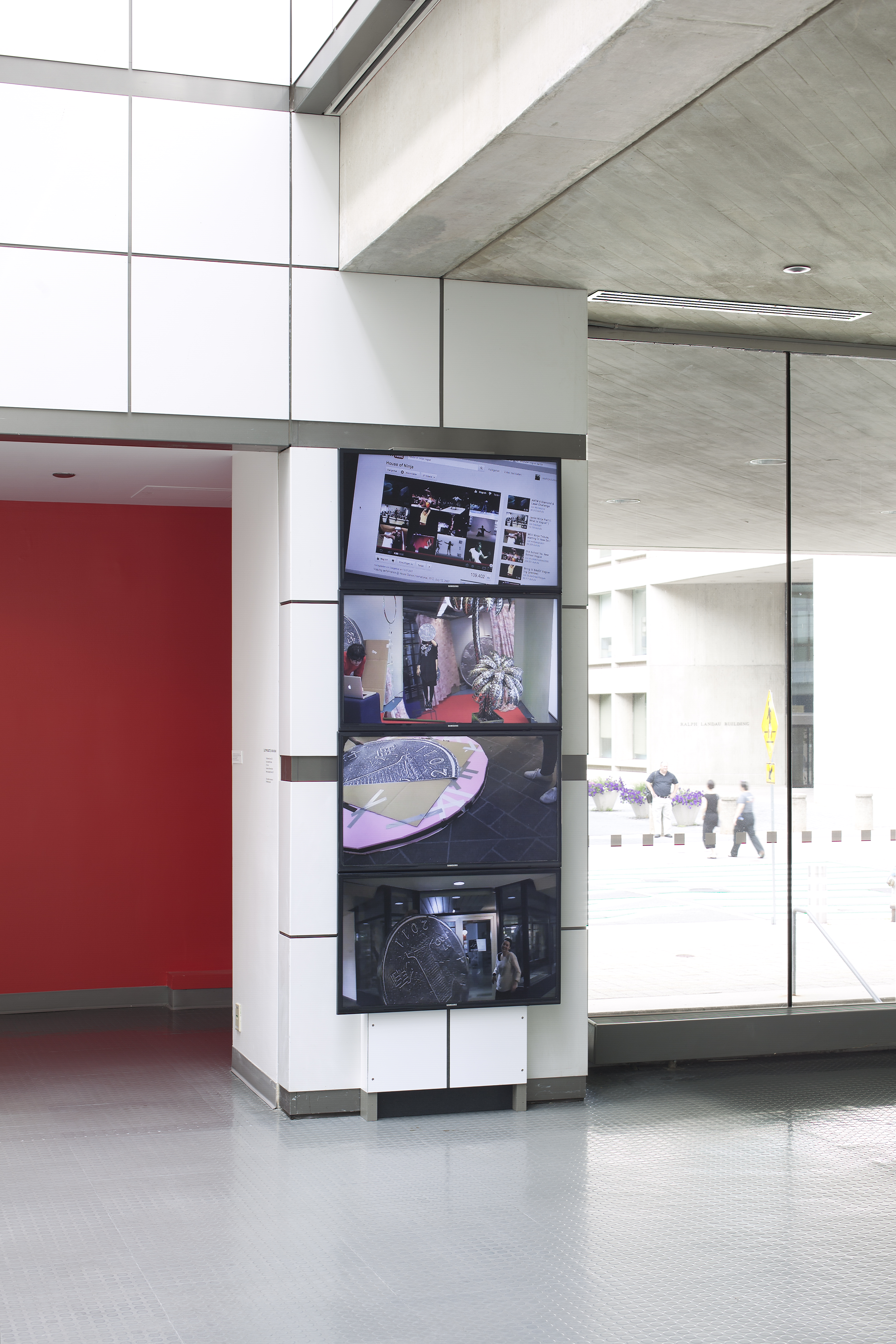 Four separate videos stacked together, one on top of the other, play on a narrow wall between an alcove and a wall of windows.