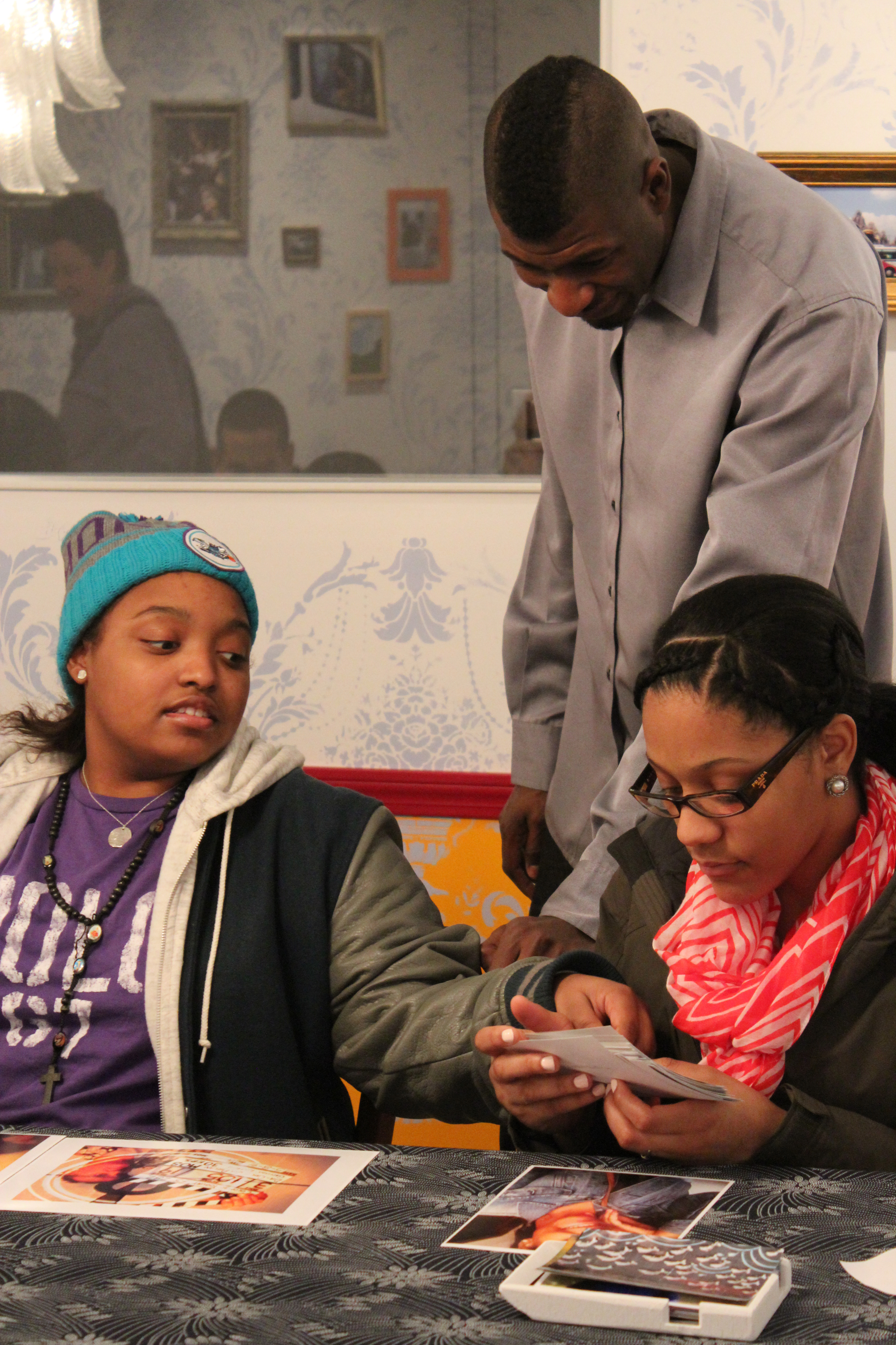 Two young women sit side by side, 1 of them points to a photo that the other holds. The artist stands above them and observes.