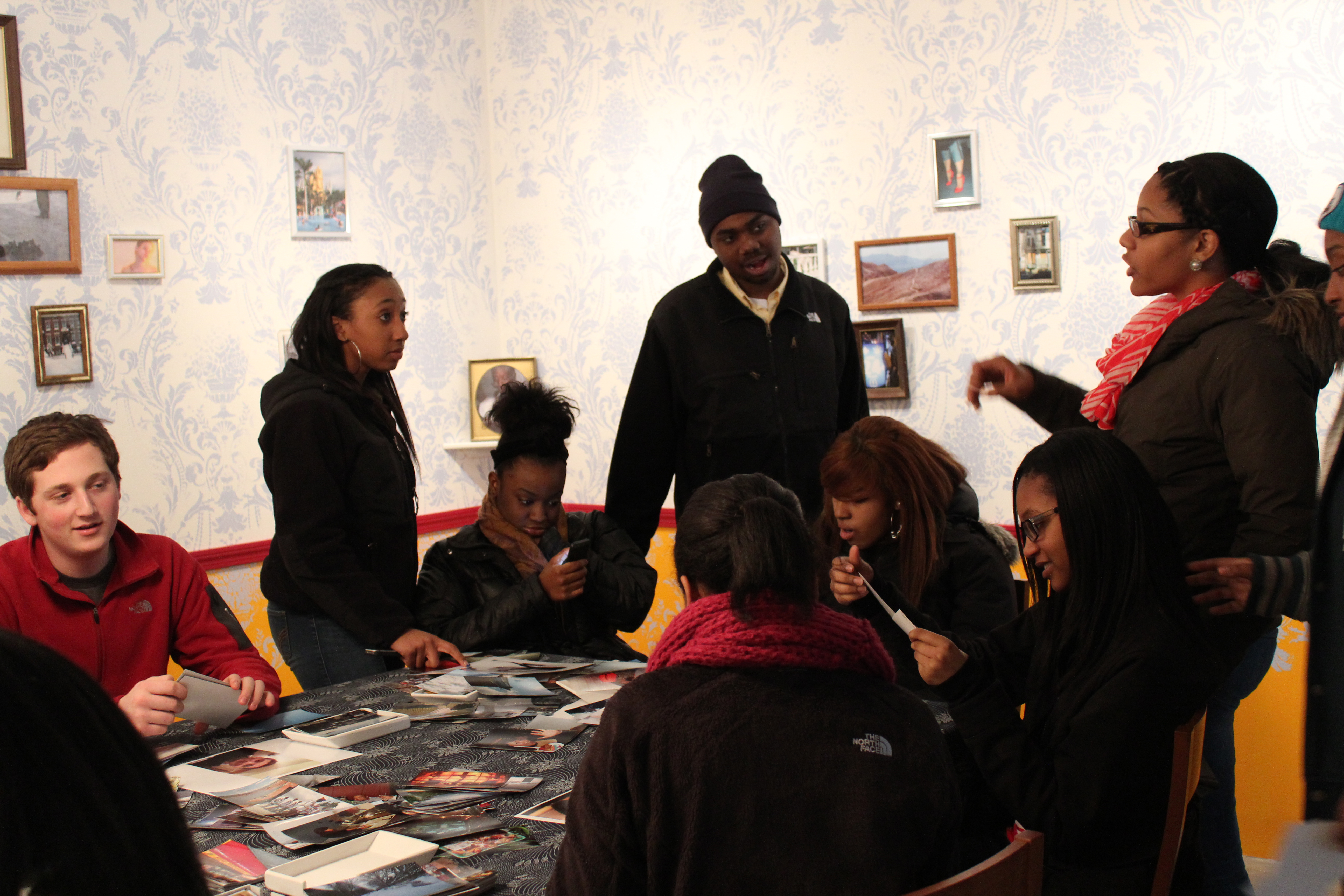 Students interact around a table filled with photos, the patterned walls behind them are dotted with small framed pictures.