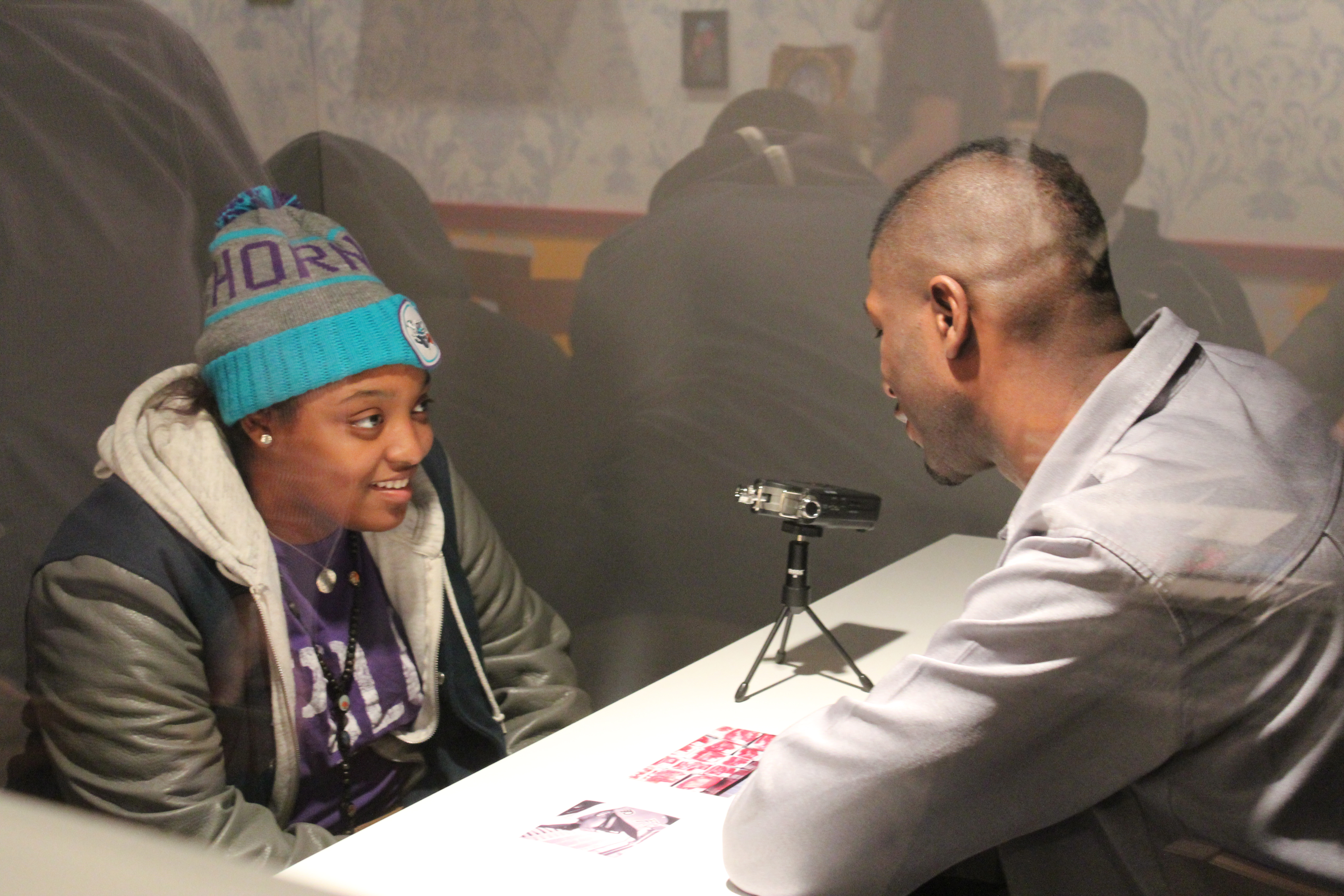 A young woman in a coat and hat sits across from a man looking down at 2 photos, a camera on a tiny tripod between them.