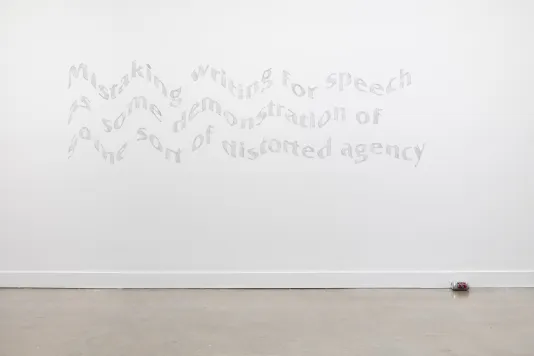 Mural featuring three lines of gray, wavy looking text on a white gallery wall with a beer can lying on its side below and slightly to the right of the mural. 