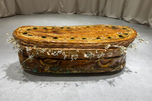A rounded wicker casket encased in resinous gelatin sits on the floor. Objects including syringes, pills, and beans adorn its top and lower sides, and dried flowers surround the opening between its lid and base.