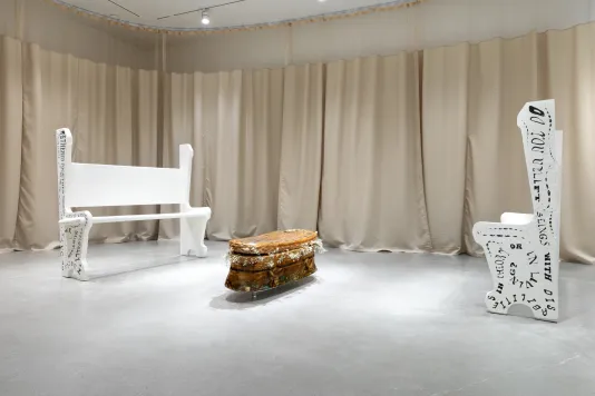 Two white church pews with text on the sides and backs frame a sculpture of a wicker casket cast in gelatin with tan hospital curtains surrounding the interior of the gallery.