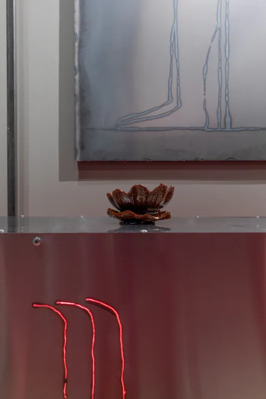 Stainless steel table with a deep red, water lily shaped incense on top, a carving of three curved lines lit red by a heating lamp on the front of the table and a welded steel drawing in the background. 