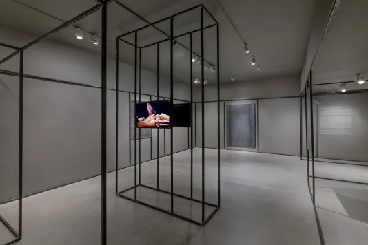 Gallery with grey walls and flooring featuring two monitors facing back-to-back on an open steel sculpture that runs throughout the gallery with two welded steel drawings in the background. 
