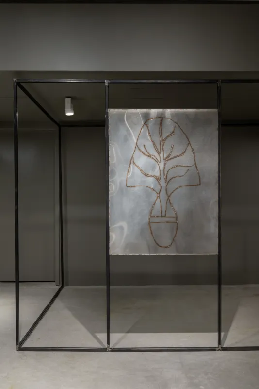 A drawing of a myrrh tree welded onto a stainless-steel panel hangs in between a large, black steel structure that forms an outline of a rectangle with vertical bars along the left and right edges of the drawing. 