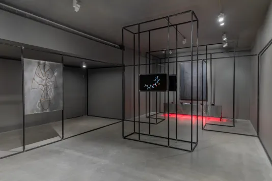 Gallery view with grey walls and cement flooring featuring a welded steel drawing on the left, a monitor in the center, a steel platform at rear glows red from heat lamps underneath, and a black steel architecture snakes along the periphery of the space. 