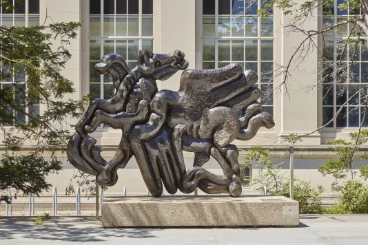 Bronze sculpture on a pedestal featuring a pegasus figure with the horse head facing backwards against a large stone building with long windows. 