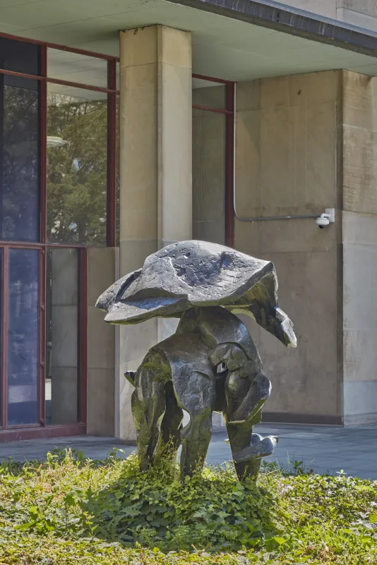Bronze sculpture resembling an abstract helmet in a small green courtyard with a building behind it. 