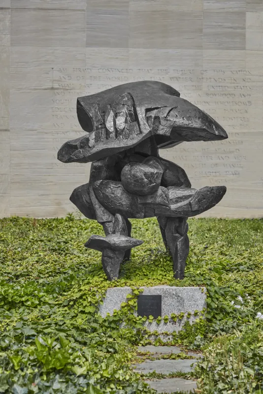 Bronze sculpture resembling an abstract helmet in a small green courtyard and a stone wall in the backhground.