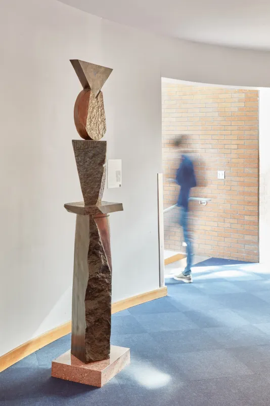 Bronze sculpture featuring different shapes stacked on top of one another and a person blurred in motion walking through a doorway in the background..