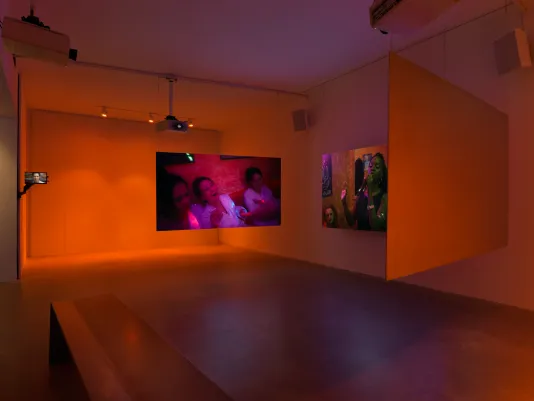 Installation view of a glowing orange gallery with three partially-obstructed suspended screens showing people laughing at a party and singing karaoke. 