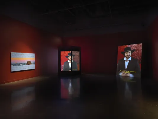 A painting of an oceanic sunset and two screens with a man in black cowboy hat and a bolo tie addressing the camera are in a dim, red-walled gallery. 