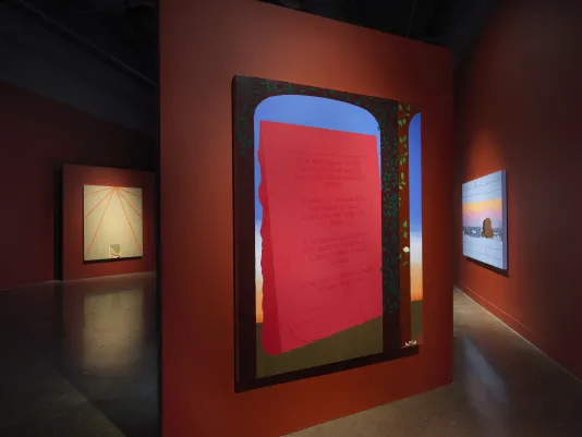 Three paintings are hung on dimly-lit red walls across a gallery.