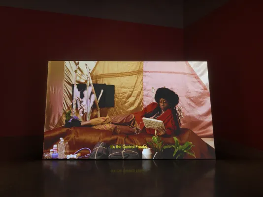 On a horizontal screen, a woman wearing a black muff hat and red jacket reclines on her side and reads a tablet in an interior draped with silks. 