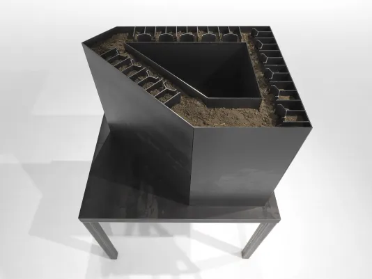 An aerial view of a dark, hollow steel sculpture with geometric inner walls is filled with dry dirt.
