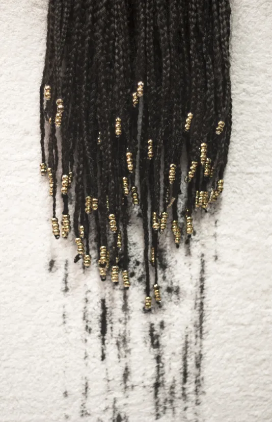 Photograph of a grouping of braided hair with small gold beads at the end of each strand. There are dark markings on the white background underneath the braids.