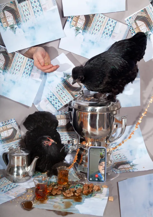 Image of two black chickens one is standing on a silver trophy the other is standing on the table. There are a collection of papers posted on the wall. There is a phone on Facetime leaning on the trophy.