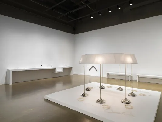 On a low pedestal in the foreground, seven standing lamps are conjoined by a single ring-shaped lampshade. A neat coil of pale cord sits near the base of each lamp. Behind it, small glass sculptures sit on a shelf, indistinct from the white wall behind them. A chevron-shaped wall piece is affixed to the wall nearby.