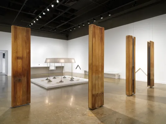 In the foreground, four oblong sculptures hang in formation, each made from planks of reclaimed wood. They hover an inch or so above the floor. Other artworks are visible in the background, including a large sculpture made of seven conjoined lamps and two chevron-shaped wall pieces.