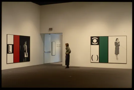 A viewer stands by the entrance to the gallery between two works, both containing figures, solid colors, and graphic symbols. 
