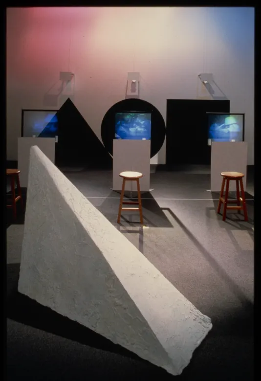 Three screens are set in front of stools. Behind them are black shapes on the wall with a prism sculpture in the foreground.