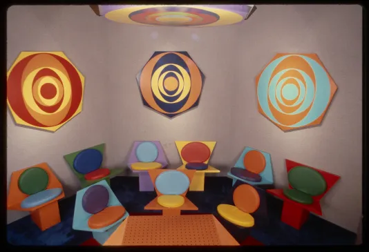 Rainbow target on ceiling above three heptogonal shapes with colorful concentric circles hanging above 2 rows of colorful seats.