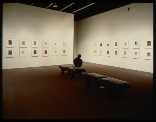 A visitor sits on gallery benches while viewing the framed works that are hung on the walls around them.  