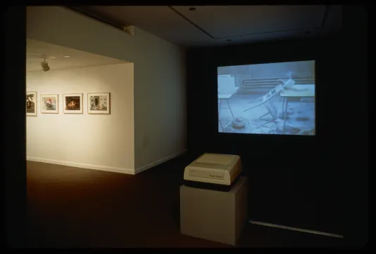 A projector shows a film in a dark gallery showing a chair resting against a table. Hanged works are lit on a wall nearby.
