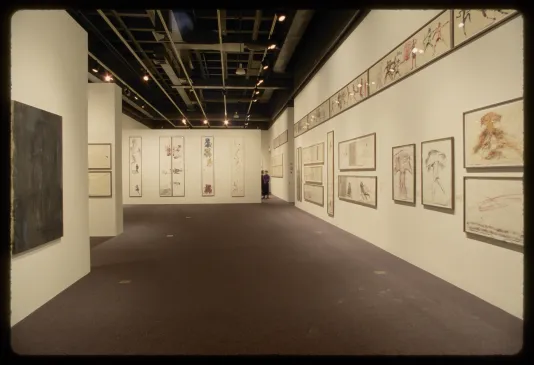 Thirty framed works on paper with colorful figurative sketches line the walls of a gallery. Two people stand in the distance.