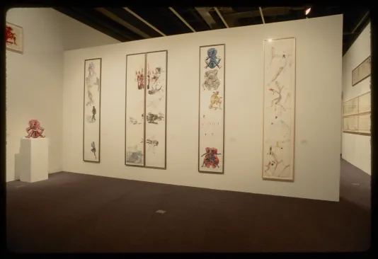 Five tall framed works on paper with colorful human figures hang adjacent to a red sculpture of a human figure on a pedestal.