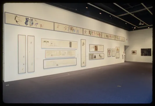 Eighteen long rectangular framed works on paper with words and colorful human figures in ancient style hang on adjacent walls.