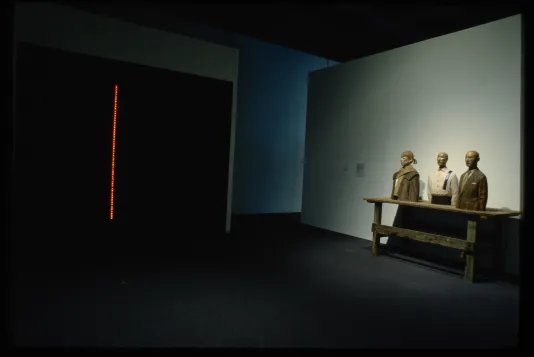 A tall strip of orange light stands out in a dark room, on the right is a sculpture of 3 people from the waist up atop a table.