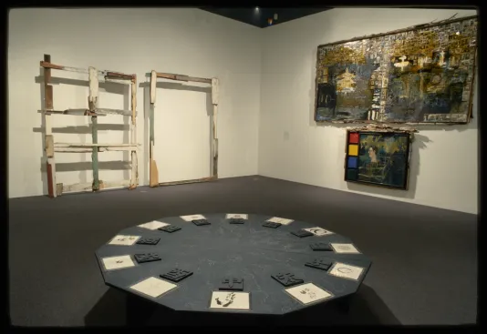 A close view of a 12 sided table in front of 2 scrap wood grid sculptures and 2 unique wood framed paintings hung behind. 