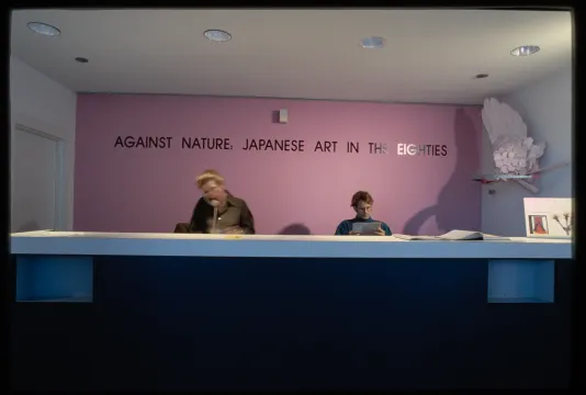 2 people sit at a desk in front of a bright pink wall with the exhibit name in black, a shelf on the right holds a sculpture.  