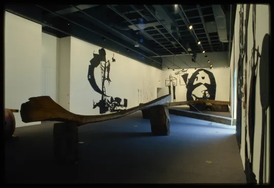 Giant wooden bench structure sits in middle of room, with black paper cut outs of abstract figures on the walls.