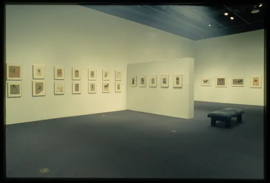 Framed drawings of figures and animals are hung on the gallery walls while a bench is placed for viewing the work.