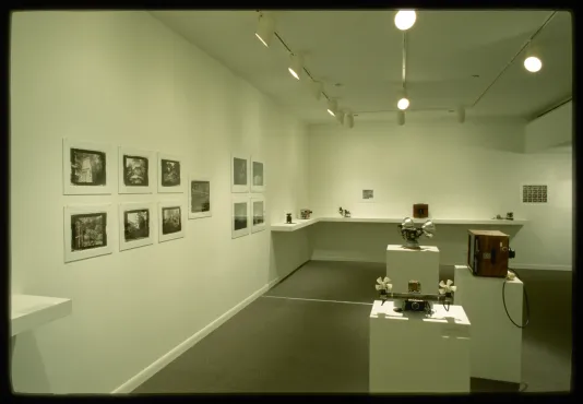 Different types of cameras sit on pedestals and on shelves. Black and white photographs are pinned to the gallery wall.