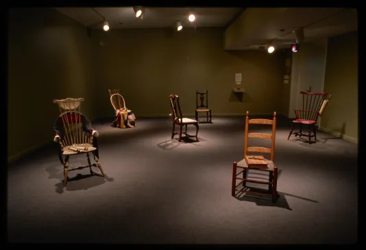 Dim room with spotlights on six spaced out colorful 17th and 18th century chairs with household items sitting on them. 