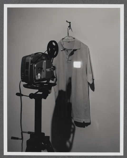 A collared shirt hangs on the gallery wall. A projector projects a tiny image onto the upper right hand side of the shirt. 