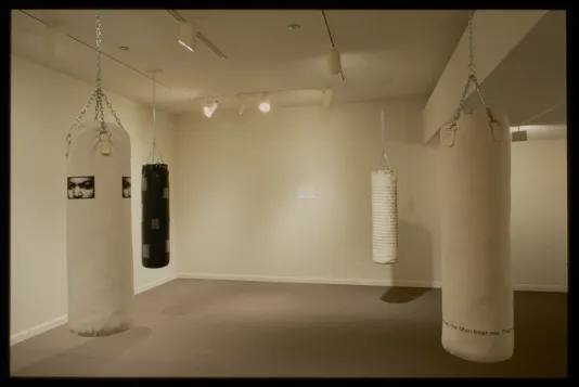 Three white and one black punching bags with text and images printed on them hang in four corners of gallery. 