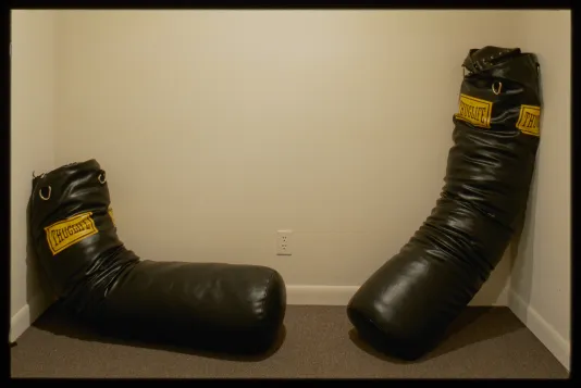 Two black punching bags with yellow logo reading “Thuglife” sit slumped across from eachother on gallery wall